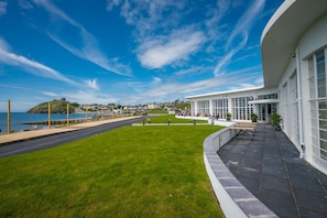 Un bâtiment moderne doté de grandes fenêtres, une zone pavée avec des bancs, et une vue sur la mer et une colline surmontée d’un château.