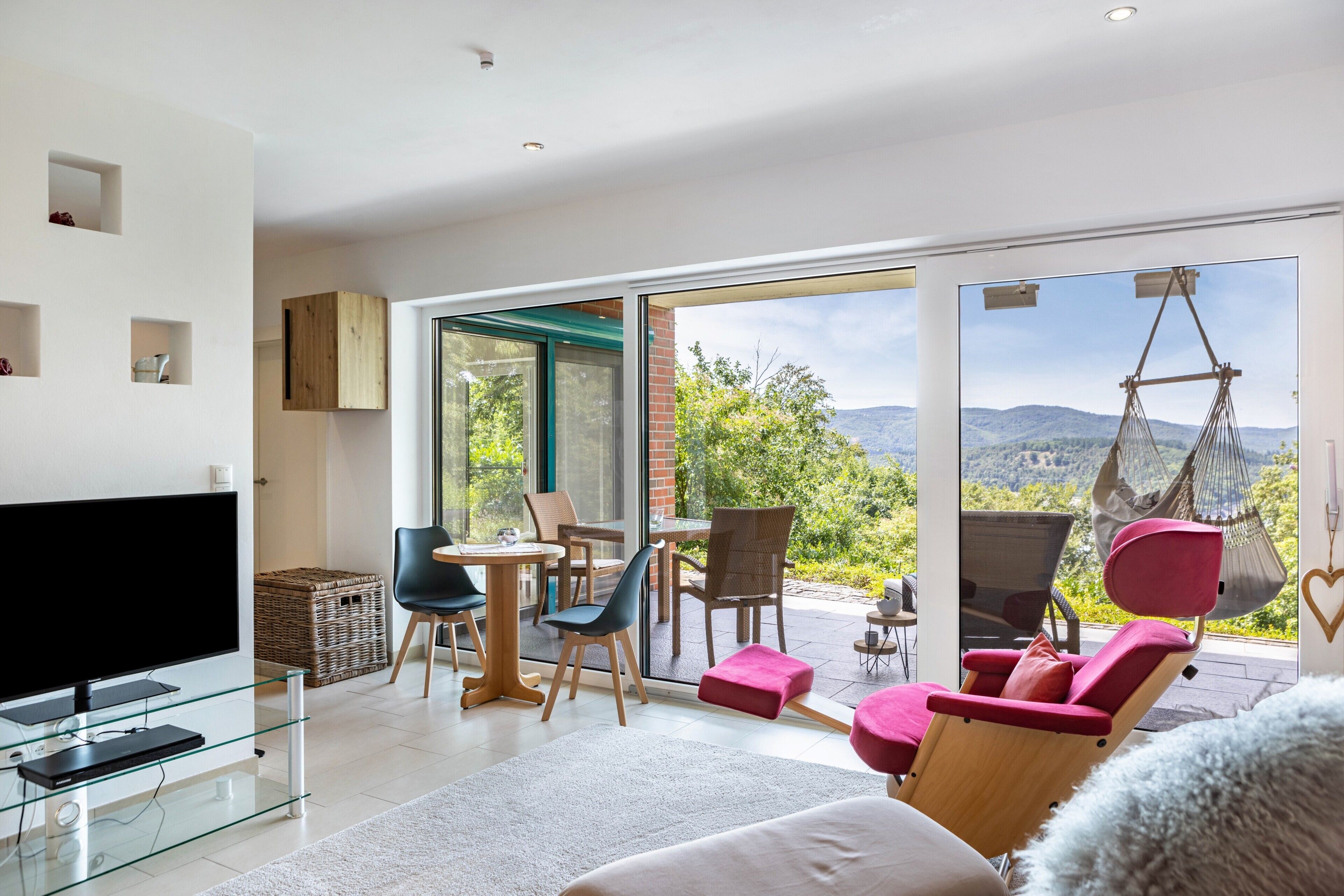 A modern living room with a flat-screen TV, glass furniture, and a view of a mountainous landscape through large sliding glass doors.