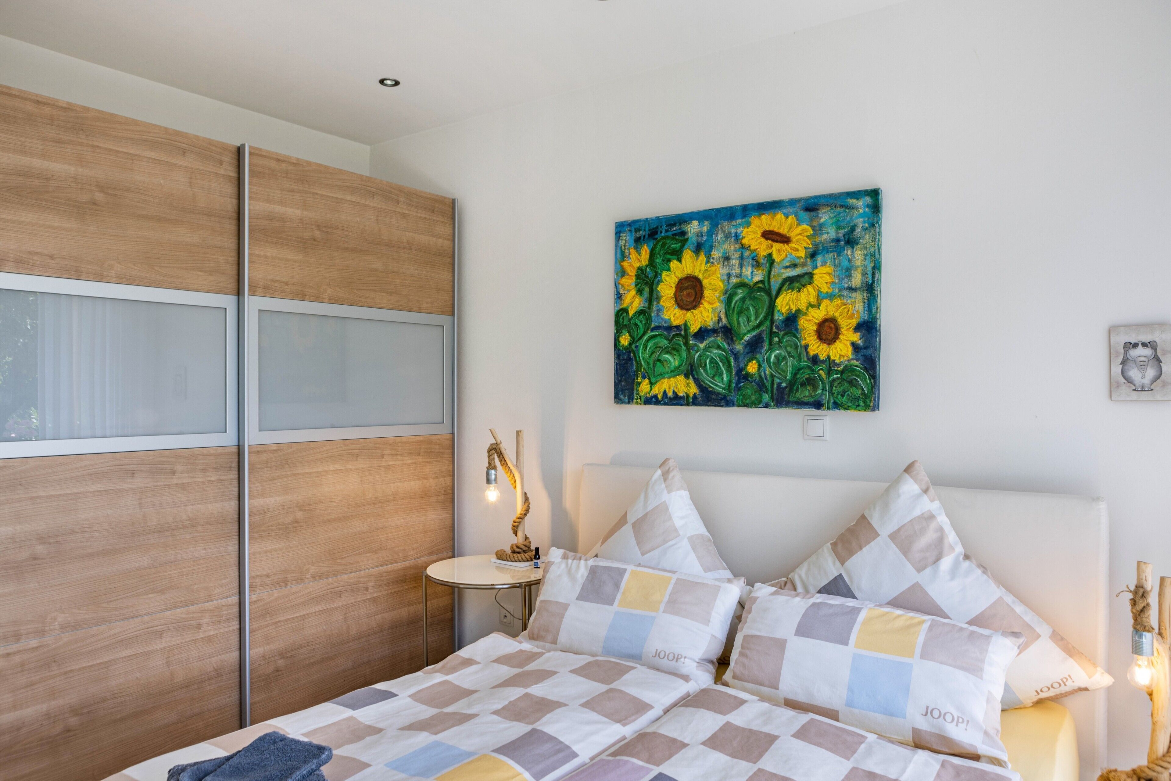 Ein Schlafzimmer mit einem Bett, einem Sonnenblumenbild, einer Nachttischlampe und einem Fenster.