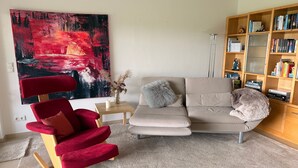 A living room with a red chair, a beige sofa, a bookshelf, and a large abstract painting.