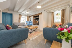 A living room with a blue sofa, a coffee table, and a TV. There is a fireplace and a window with curtains. - Beautiful Cosy Cottage Overlooking Beaumaris Bay | Rallt-Goch-Bach (Beaumaris)
