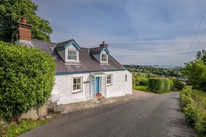 A white cottage with a blue door and a small porch, surrounded by greenery and a gravel driveway. - Beautiful Cosy Cottage Overlooking Beaumaris Bay | Rallt-Goch-Bach (Beaumaris)