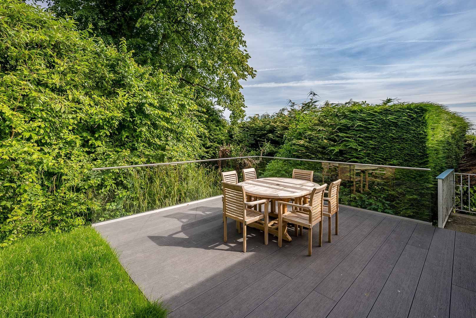 A wooden deck with a round table and chairs, surrounded by lush greenery.