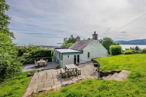 A white cottage with a blue roof, a patio with a table and chairs, and a view of the sea. - Beautiful Cosy Cottage Overlooking Beaumaris Bay | Rallt-Goch-Bach (Beaumaris)