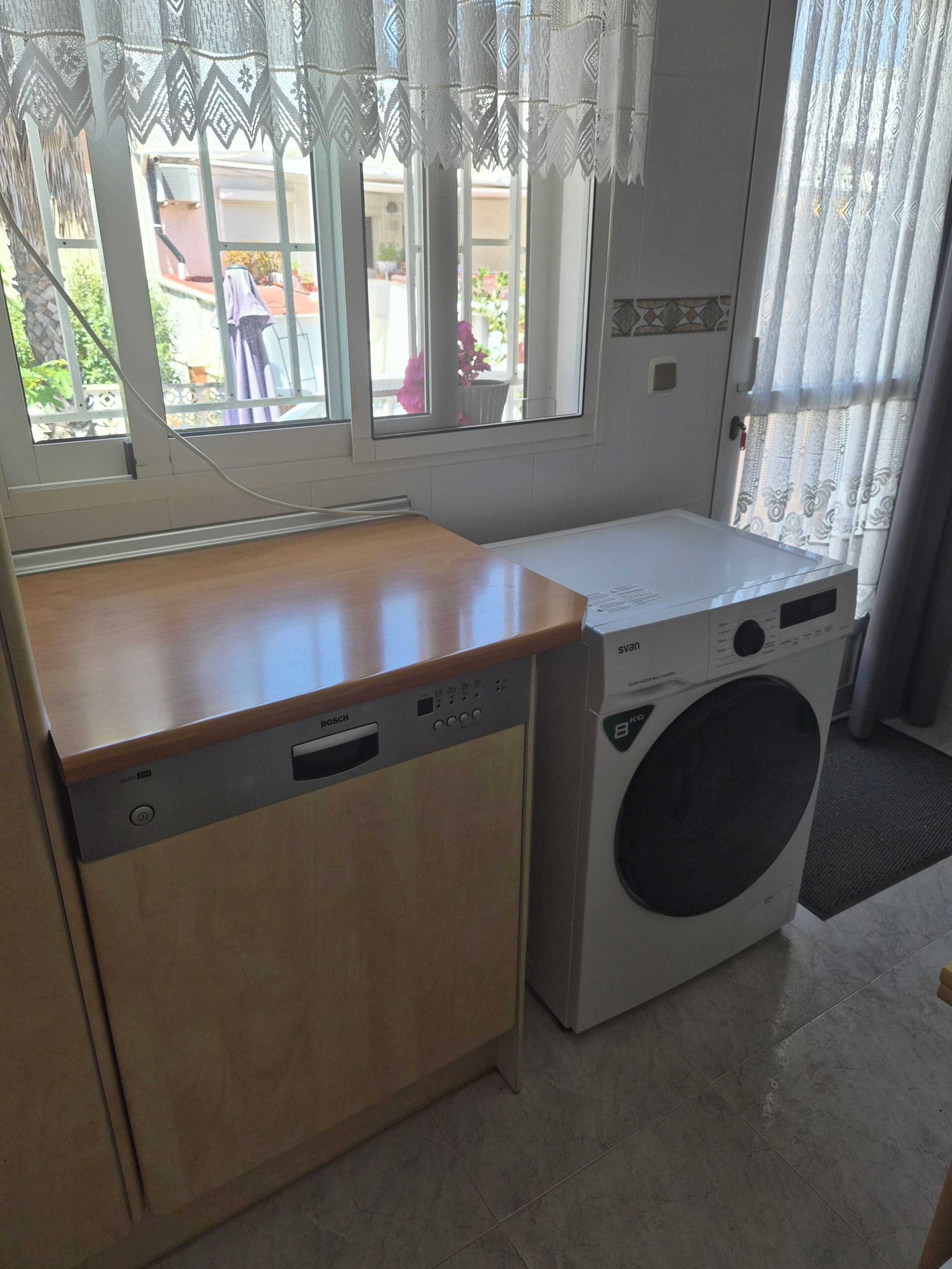 Una cocina compacta con lavavajillas Bosch y lavadora LG, una ventana con cortinas de encaje y vistas al exterior.