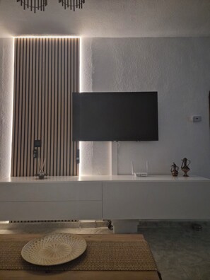 A modern living room with a flat-screen TV mounted on a white wall, a textured wall panel, a small table with a decorative plate, and a woven mat. - Casa Luginilla- am Meer-85m²-mit großem Pool (Cartagena)