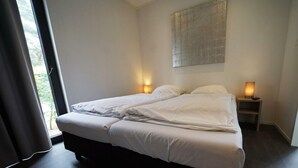 A double bed with white linens, two bedside tables with lamps, a large window with curtains, and a textured wall art piece. - Holiday Home in Otterlo With Terrace (Otterlo)