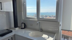 Eine Küche mit weißer Arbeitsplatte, einem Wasserkocher, einer Spüle und einem Fenster mit Blick auf eine Stadtlandschaft.