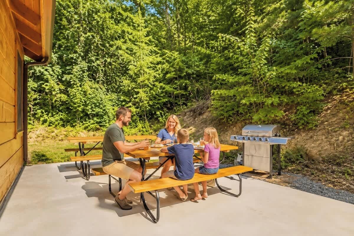 A family dining at an outdoor wooden table with a grill in a forested area.