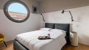 Ein Schlafzimmer mit rundem Fenster, einem Bett mit weißer Bettwäsche, einem Nachttisch mit Lampe und einem Tablett mit Tassen und einer Kanne.
