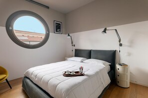 Un dormitorio con ventanal redondo, cama con ropa de cama blanca, mesita de noche con lámpara y bandeja con tazas y jarra.