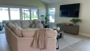 A living room with a beige sectional sofa, a flat-screen TV mounted on the wall, a wooden cabinet, and a large window with a view of the outside. - Open the Doors to Paradise in This Stunning New Retreat! (Sumterville)