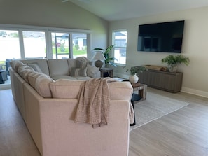 A living room with a beige sectional sofa, a flat-screen TV mounted on the wall, a wooden cabinet, and a large window with a view of the outside. - Open the Doors to Paradise in This Stunning New Retreat! (Sumterville)