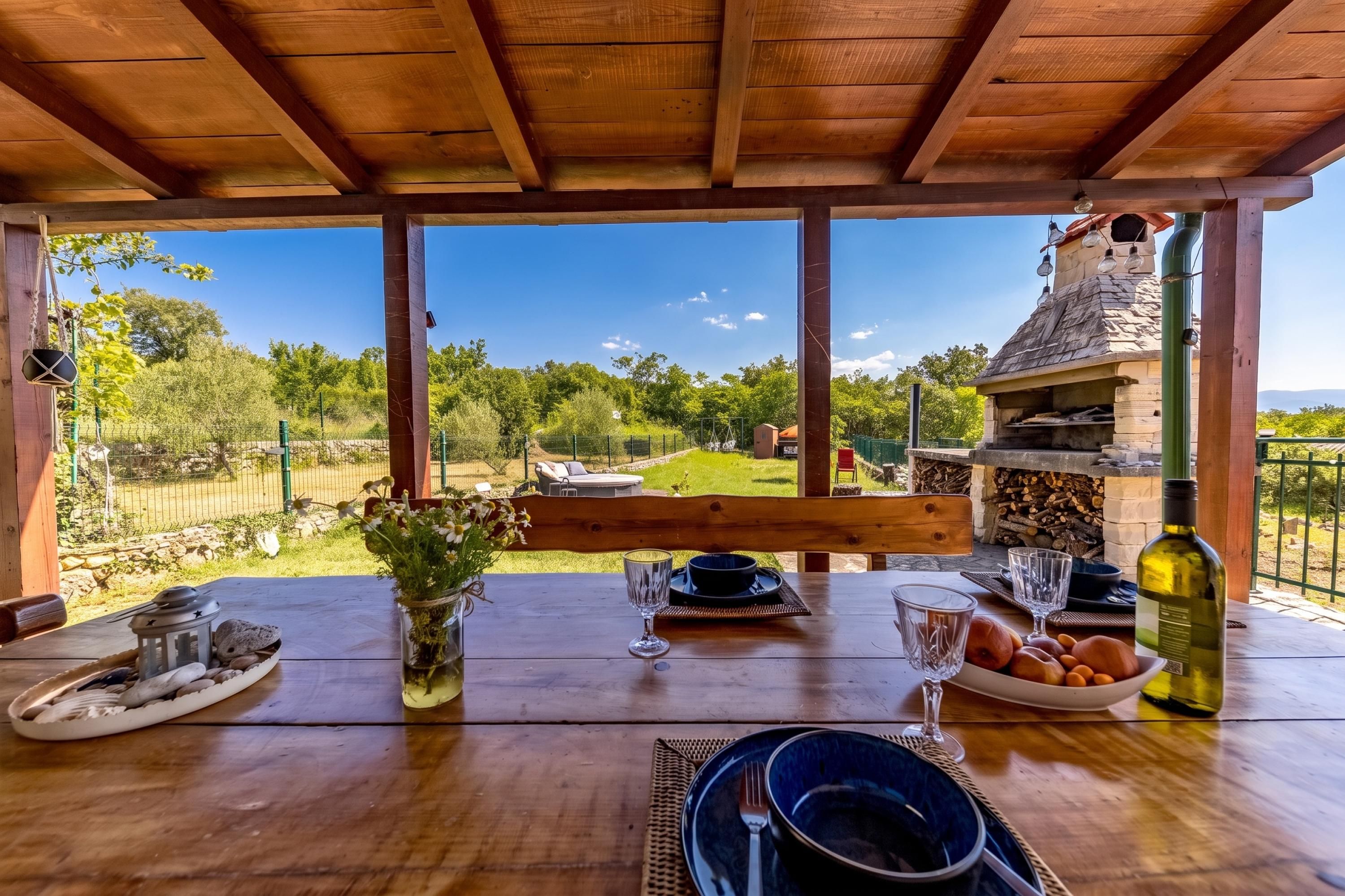 Un'area pranzo all'aperto con tavolo in legno, forno a pietra e vista su giardino e alberi.