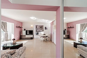 Ein modernes Wohnzimmer mit einer Couch, einem Essbereich mit Stühlen, einer Küche inklusive Fernseher und einem Balkon mit Vorhängen.