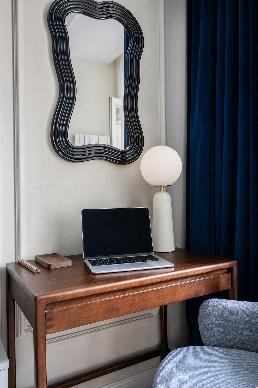 A laptop on a wooden desk with a mirror above it, a lamp beside the desk, and a blue curtain in the background.