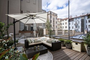 Eine Dachterrasse mit einer Eckcouch, einem Glastisch, Topfpflanzen und einem großen Sonnenschirm.