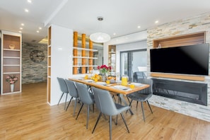 Ein moderner Essbereich mit einem Holztisch, grauen Stühlen, einer Deckenleuchte und einem an der Wand montierten Fernseher.