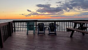 A wooden deck with Adirondack chairs and a picnic table overlooking the ocean at sunset.
