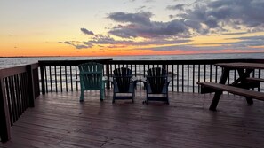 Eine Holzterrasse mit Adirondack-Stühlen und einem Picknicktisch mit Blick auf den Ozean bei Sonnenuntergang.