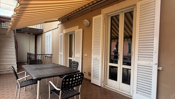 Uma área de refeições ao ar livre com uma mesa e cadeiras, um toldo listrado e persianas brancas nas paredes.