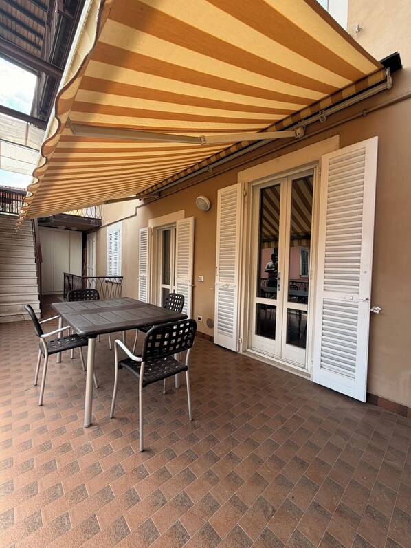 Ein Essbereich im Freien mit Tisch und Stühlen, einem gestreiften Sonnendach und weißen Fensterläden.