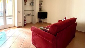 Uma sala de estar com um sofá vermelho, uma televisão de tela plana, uma estante de livros e uma porta que dá para o exterior.