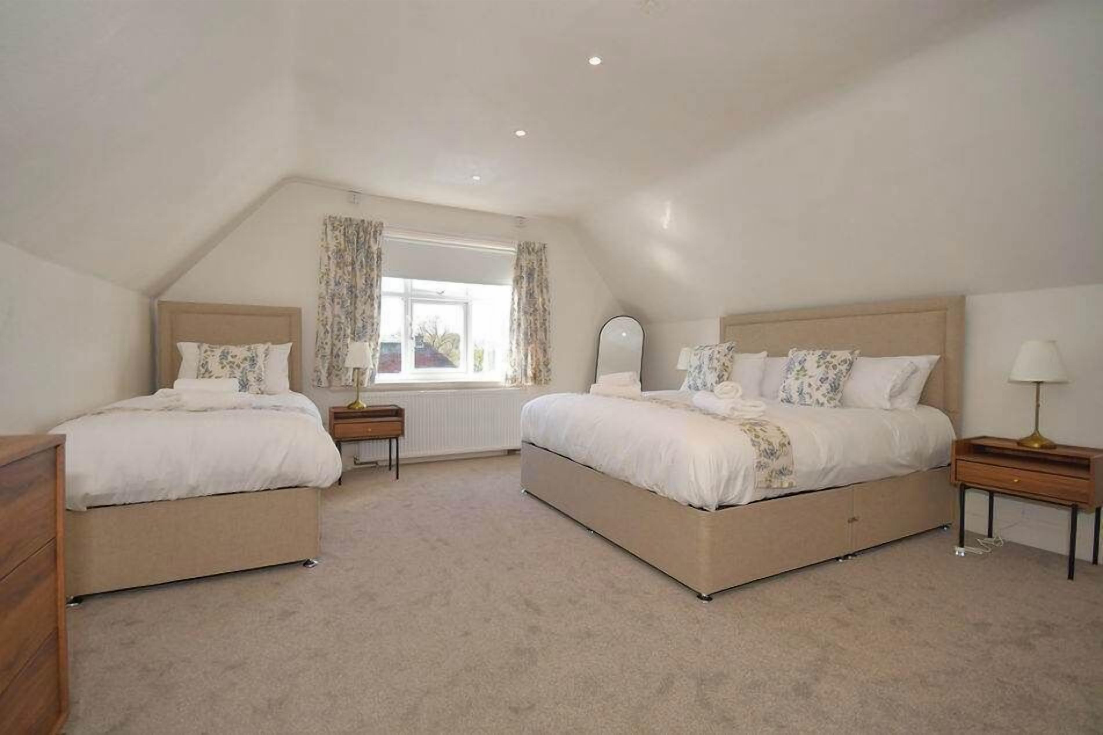 A bedroom with two beds, a window with curtains, a radiator, and a bedside table with a lamp.