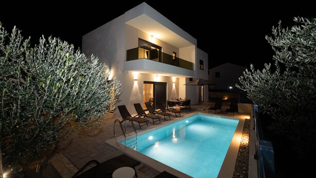 Una casa moderna de dos pisos con piscina, mobiliario de exterior y olivos.