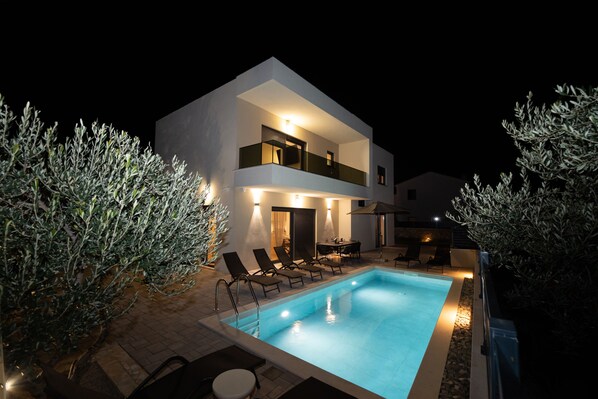Ein modernes, zweistöckiges Haus mit Swimmingpool, Sitzgelegenheiten im Freien und Olivenbäumen.