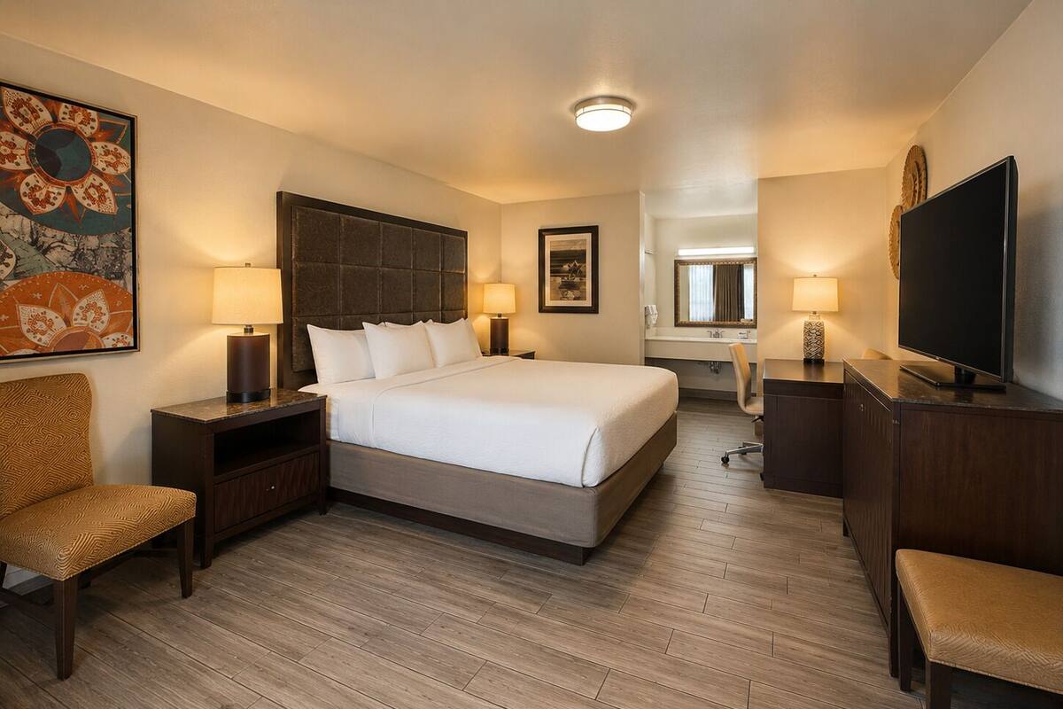 Ein Hotelzimmer mit einem Bett, zwei Nachttischen mit Lampen, einem Sessel, einem Schreibtisch mit Fernseher und einem großen Spiegel.