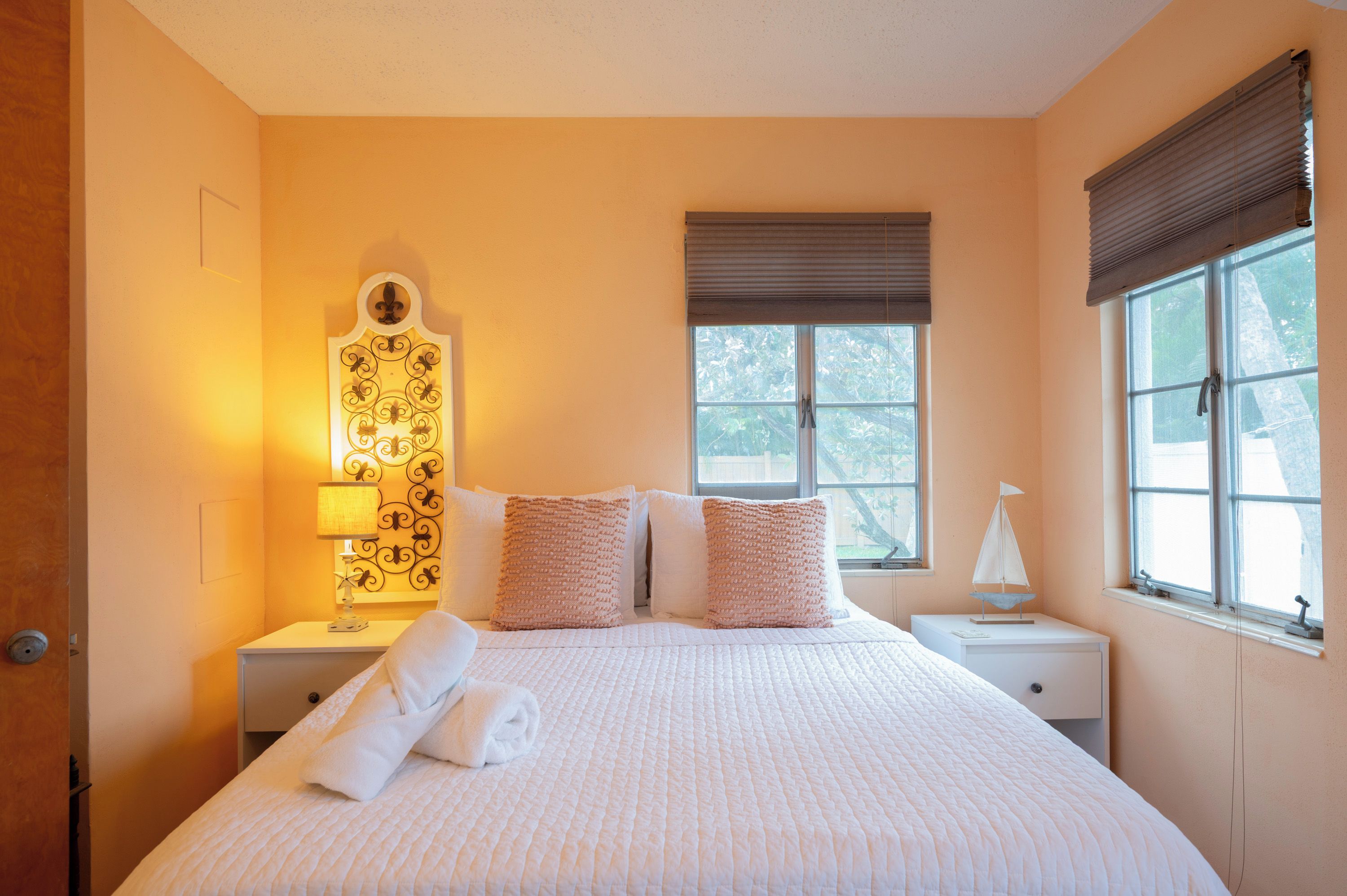 A neatly made bed with pillows and a rolled towel, a bedside lamp, a decorative wall piece, and a window with blinds.