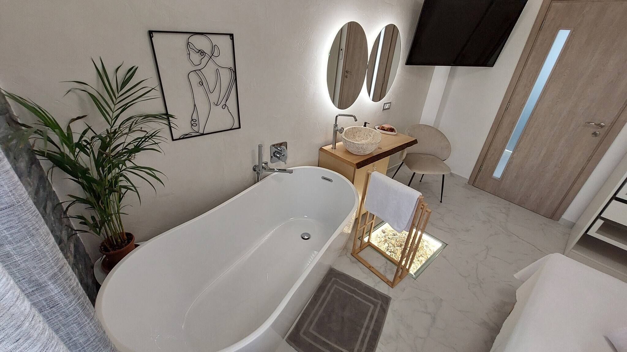 A modern bathroom with a freestanding bathtub, a wooden side table with a bowl, a chair, and a potted plant.