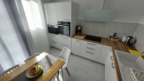 A modern kitchen with white cabinets, a built-in oven, a coffee maker, and a dining area with a table and chairs. - My Hersonissos Home - Luxury Suite (Hersonissos)