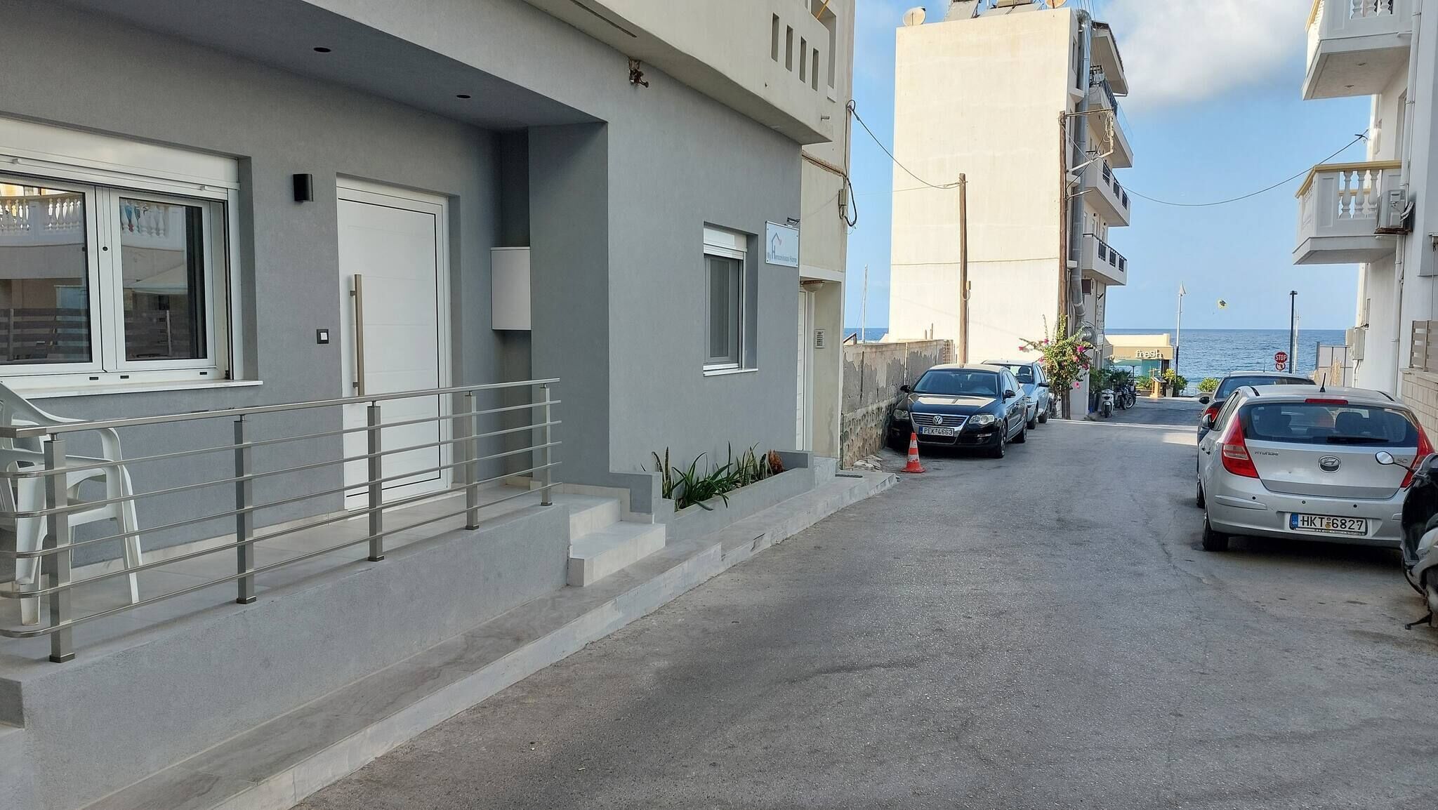A narrow street with parked cars, a white building with a balcony, and a view of the sea.