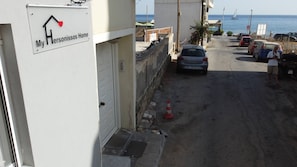 Eine schmale Straße mit einem geparkten Auto, einem Gebäude mit der Aufschrift 'My Hersonissos Home' und einem Blick auf das Meer im Hintergrund.