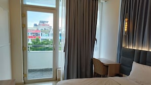 A hotel room with a bed, a desk, a chair, a window with curtains, and a view of buildings outside. - the royal oak sec 40 hotel gurgaon (Gurugram)