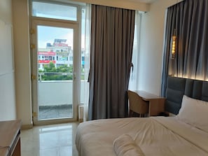 Ein Hotelzimmer mit einem Bett, einem Schreibtisch, einem Stuhl, einem Fenster mit Vorhängen und Blick auf Gebäude.