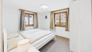 Une chambre à coucher avec un lit, une table de chevet, une lampe et deux fenêtres avec des rideaux.
