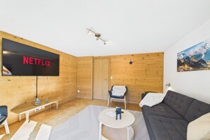 Ein gemütliches Zimmer mit Flachbildfernseher, Holzwänden, einer Couch, einem Sessel und einem kleinen Tisch.