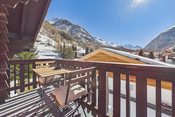 Un balcón de madera con una mesa y una silla, con vistas a un paisaje montañoso nevado.