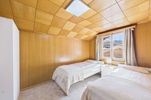 Une chambre avec des murs lambrissés, un plafond en pente, deux lits, une table de chevet, une fenêtre avec des rideaux et une vue sur un paysage enneigé.