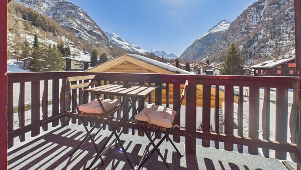 A wooden deck with a table and chairs, overlooking a mountainous landscape with snow-covered peaks and a clear sky.