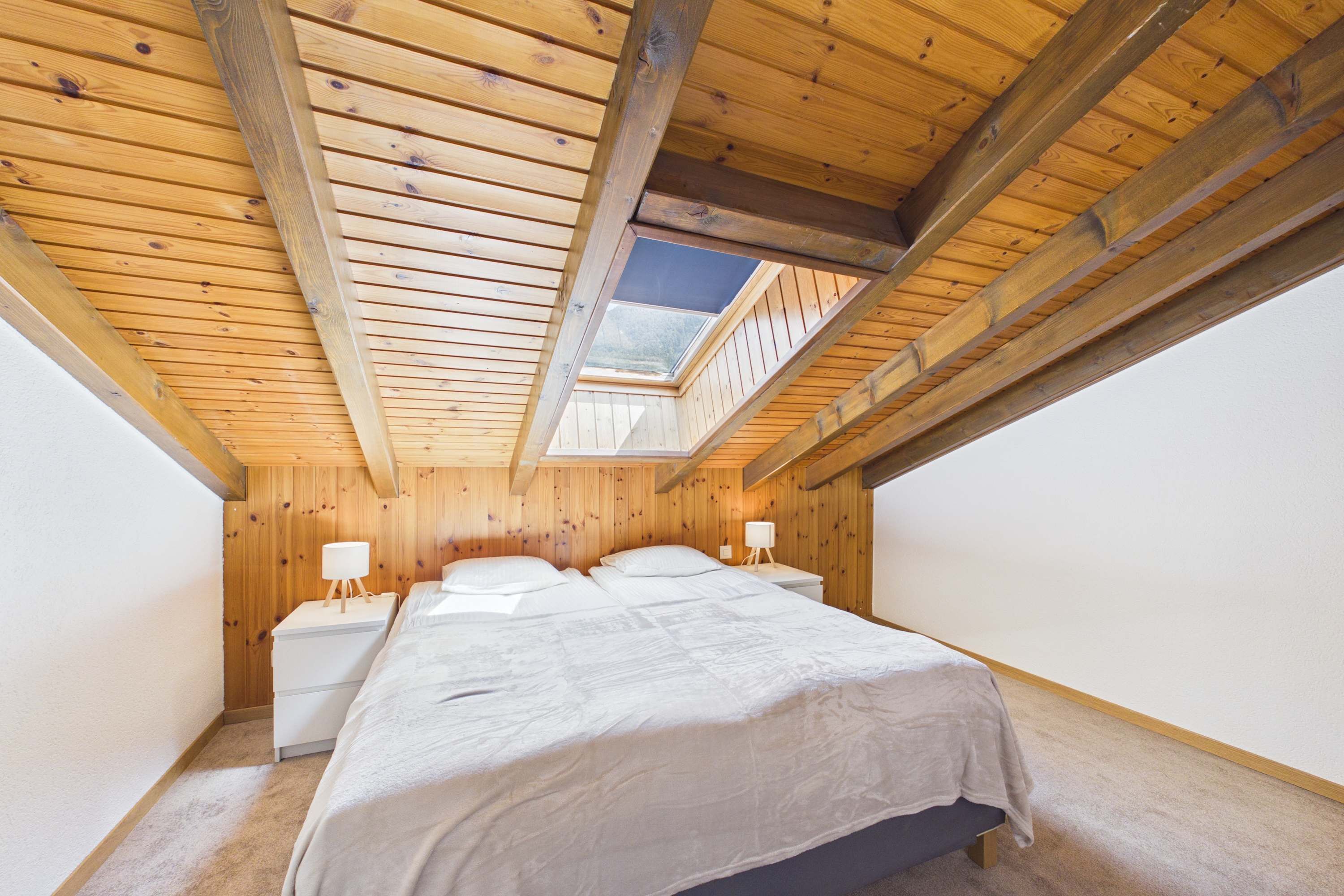 A bedroom with a sloped ceiling, wooden beams, a skylight, a bed, bedside tables, and a nightstand.