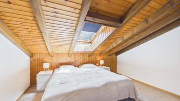 A bedroom with a sloped ceiling, wooden beams, a skylight, a bed, bedside tables, and a nightstand.