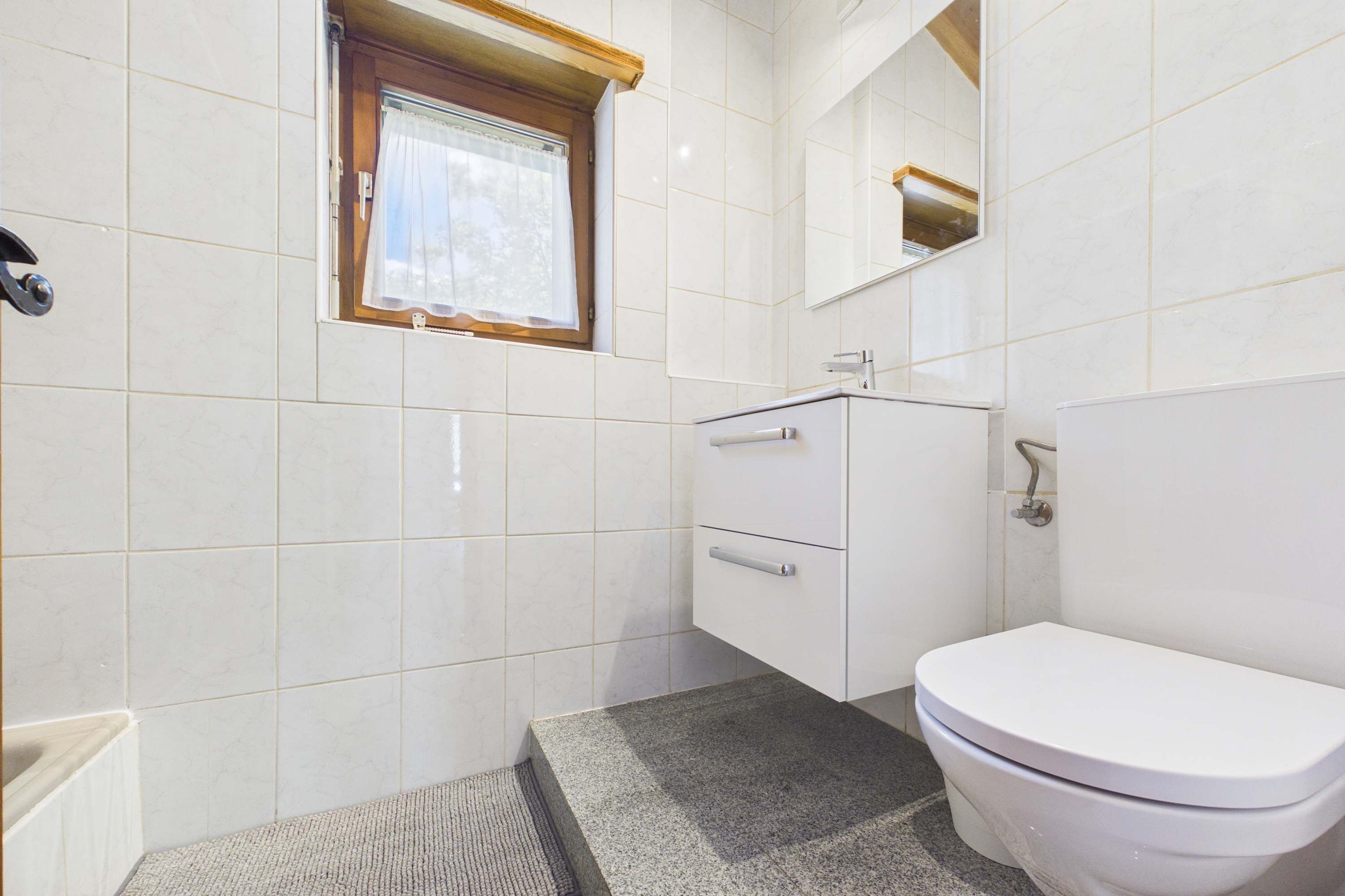 A bathroom with white tiles, a window with a curtain, a sink, a toilet, and a mirror.