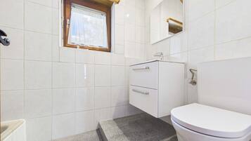 A bathroom with white tiles, a window with a curtain, a sink, a toilet, and a mirror.