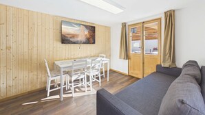 A room with a wooden wall, a flat-screen TV, a dining table with chairs, a gray sofa, and a sliding glass door.