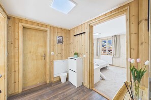 Una habitación con paredes de madera, una cama, un escritorio, un armario blanco pequeño, una ventana con cortinas y un tragaluz.