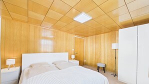A bedroom with a wooden ceiling, a large bed, a nightstand with a lamp, a small stool, and a wardrobe.